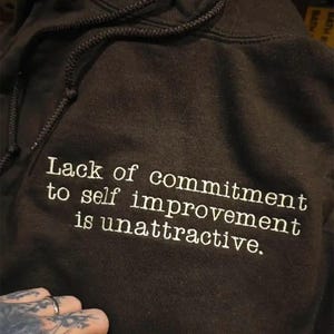 Dark Chocolate hoodie with white embroidered quote reading “Lack of commitment to self improvement is unattractive.” The text is stitched in a clean typewriter-style font across the chest, creating a bold minimalist statement. High-contrast white thread on black fabric emphasizes the motivational quote design.