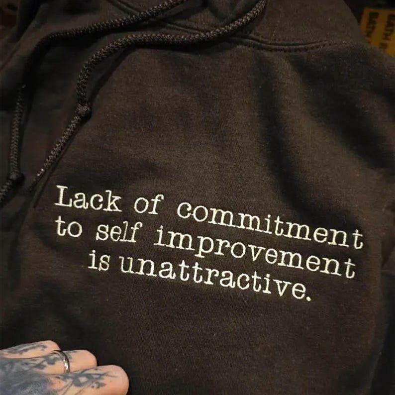 Dark Chocolate hoodie with white embroidered quote reading “Lack of commitment to self improvement is unattractive.” The text is stitched in a clean typewriter-style font across the chest, creating a bold minimalist statement. High-contrast white thread on black fabric emphasizes the motivational quote design.
