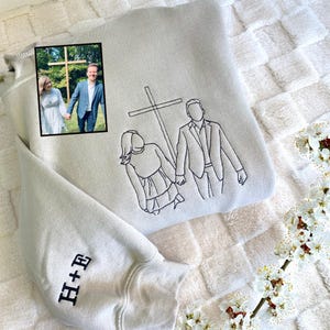 May include: A light gray hoodie with a black embroidered outline of a couple holding hands, with a cross in the background. A small framed photo is on the hoodie. The sleeve has the embroidered initials "H+E".