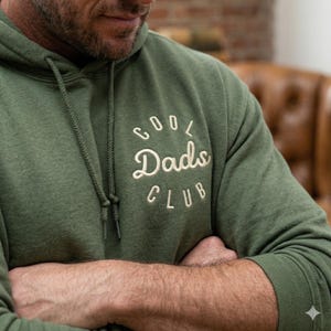 May include: Olive green hoodie with the embroidered text "Cool Dads Club" in a white, cursive font. The hoodie has a drawstring hood and a kangaroo pocket. The person in the image has their arms crossed.