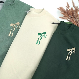 Può includere: Tre felpe girocollo nelle tonalità del verde e del crema. Ogni felpa presenta un fiocco ricamato con una singola iniziale. Le felpe sono realizzate in un materiale morbido, perfette per l'abbigliamento casual.