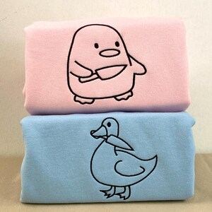 May include: Two folded, soft fabric items, one pink and one blue, stacked on top of each other. The pink item features a cartoon penguin holding knives, while the blue item has a cartoon duck. Both designs are outlined in black.