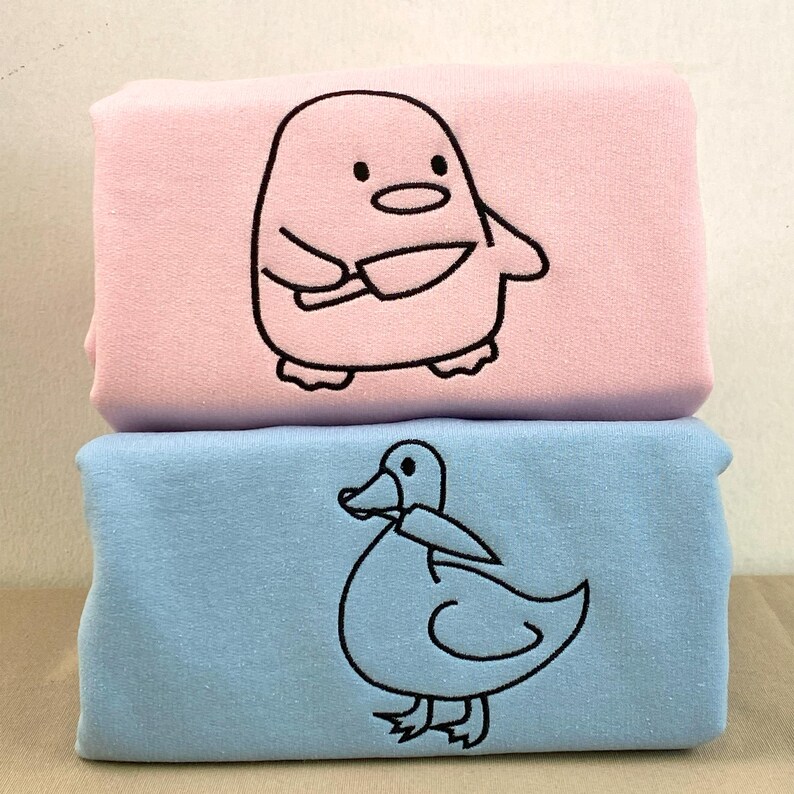 May include: Two folded, soft fabric items, one pink and one blue, stacked on top of each other. The pink item features a cartoon penguin holding knives, while the blue item has a cartoon duck. Both designs are outlined in black.