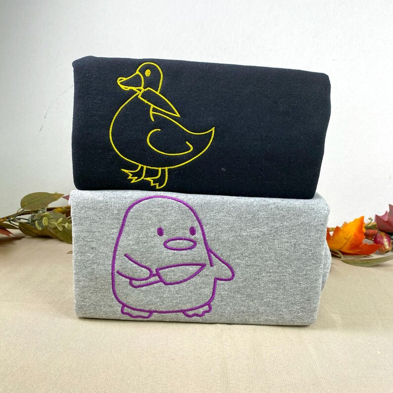 May include: Two folded sweatshirts, one black with a yellow duck outline and one gray with a purple penguin outline. The duck and penguin are embroidered on the front of the shirts. The sweatshirts are stacked on top of each other.