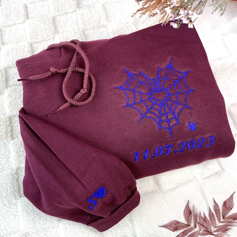 Personalized maroon hoodie with blue embroidered spider web heart design, custom anniversary date "11.07.2023" on the chest, and matching initial with heart on the sleeve. Unique romantic Halloween couple sweatshirt, custom embroidered gift for anniversary or spooky season outfit.