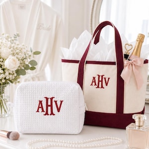May include: A white waffle-textured cosmetic bag and a cream-colored tote bag with burgundy accents, both embroidered with the letters AHV. The tote bag contains tissue paper, a champagne bottle, and a heart-shaped bottle opener. A white robe with "Bride" embroidered on it hangs in the background.