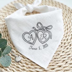 May include: White bandana with embroidered gray hearts, initials "A" and "R", and the date "June 5, 2025". The bandana is tied at the top and rests on a woven surface. A silver locket is visible in the lower left corner.