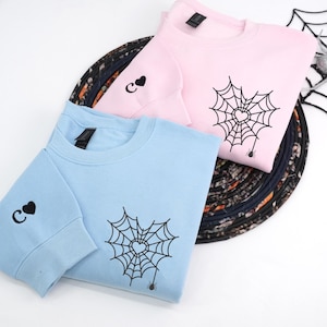 Light pink and light blue sweatshirts displayed in a flat lay style, featuring a spider web heart embroidery on the front chest with a small hanging spider. Sleeves include a personalized initial letter with a heart symbol embroidered near the cuff. Minimalist embroidered couple hoodies, soft pastel tones, romantic matching sweatshirt gift.
