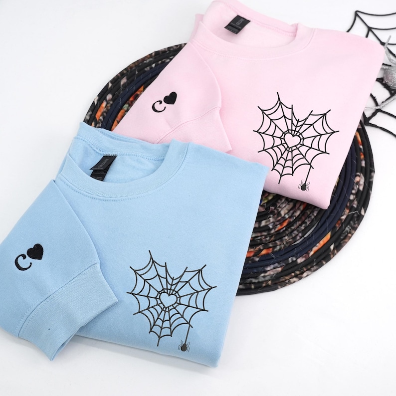 Light pink and light blue sweatshirts displayed in a flat lay style, featuring a spider web heart embroidery on the front chest with a small hanging spider. Sleeves include a personalized initial letter with a heart symbol embroidered near the cuff. Minimalist embroidered couple hoodies, soft pastel tones, romantic matching sweatshirt gift.