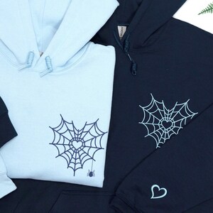 Custom embroidered Halloween couple hoodies featuring spider web with heart design on the chest and mini heart on sleeve. Matching light blue and navy blue sweatshirts, perfect spooky season outfit or gift for couples. Unique romantic gothic embroidery, cozy and stylish for fall or October vibes.
