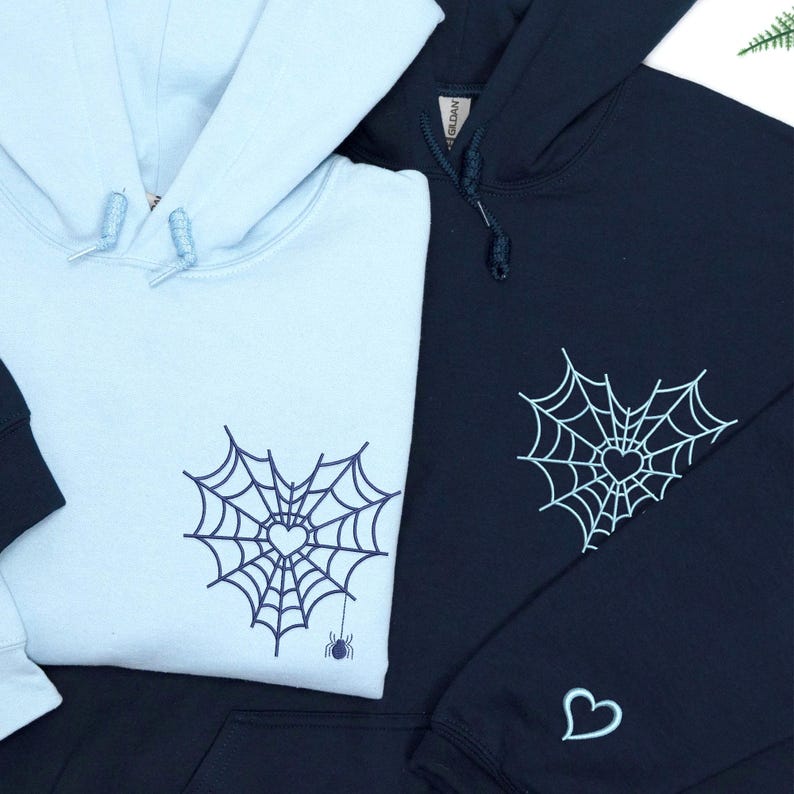 Custom embroidered Halloween couple hoodies featuring spider web with heart design on the chest and mini heart on sleeve. Matching light blue and navy blue sweatshirts, perfect spooky season outfit or gift for couples. Unique romantic gothic embroidery, cozy and stylish for fall or October vibes.