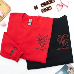 May include: Red and black sweatshirts featuring spiderweb and heart designs. The red sweatshirt has initials on the sleeve. The sweatshirts have the Roman numeral date IX.XVI.MMXXIV.