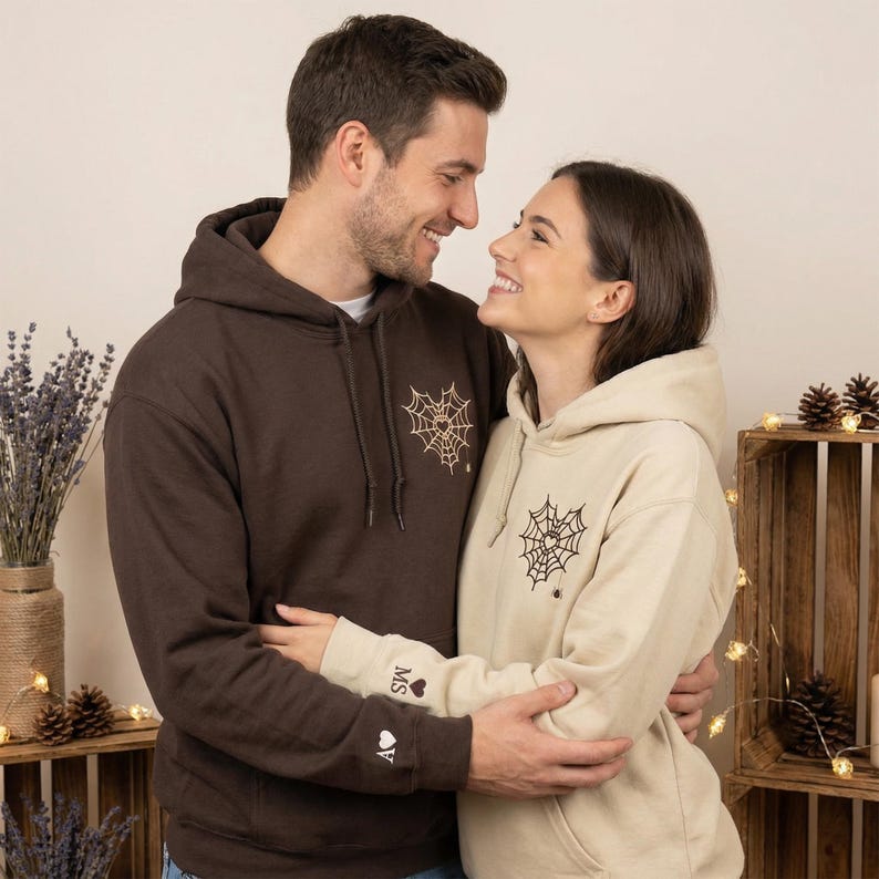 May include: Two hooded sweatshirts, one brown and one beige, feature a black spiderweb design on the chest. The brown sweatshirt has the initials "MS" and a heart with the letter "A" embroidered on the sleeve. The couple is embracing.