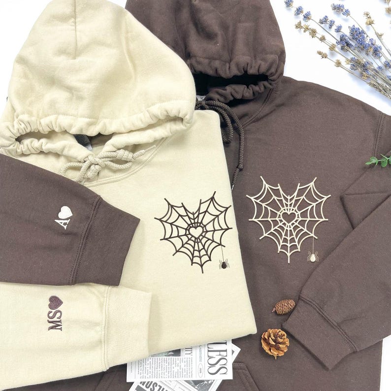 Couple Halloween hoodies with heart spiderweb embroidery and mini spider detail. Custom initials on sleeves with heart symbol. Cozy beige and brown matching sweatshirts—perfect spooky season gift for couples, best friends, or fall lovers. Unique embroidered gothic streetwear for Halloween vibes.