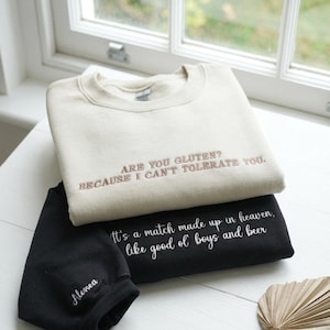 May include: Two sweatshirts, one cream-colored and one black, are stacked. The cream sweatshirt has the embroidered text "ARE YOU GLUTEN? BECAUSE I CAN'T TOLERATE YOU." The black sweatshirt says "It's a match made up in heaven, like good ol' boys and beer."