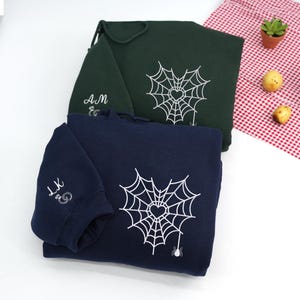 Navy and Forest Green hoodies folded on a white background, featuring minimalist spider web heart embroidery on the chest. The web design includes a small heart at the center and a dangling spider detail. Sleeve embroidery shows personalized initials in cursive script. Clean flat lay product photo, custom embroidered hoodie, cozy aesthetic gift for couples.