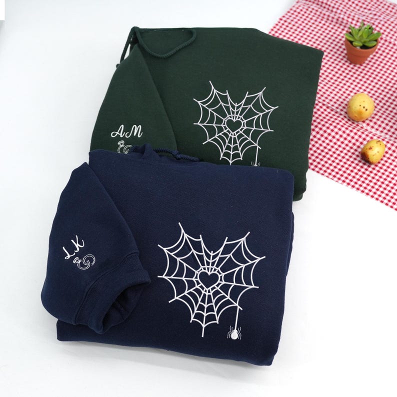 Navy and Forest Green hoodies folded on a white background, featuring minimalist spider web heart embroidery on the chest. The web design includes a small heart at the center and a dangling spider detail. Sleeve embroidery shows personalized initials in cursive script. Clean flat lay product photo, custom embroidered hoodie, cozy aesthetic gift for couples.