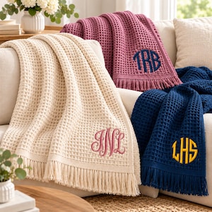 May include: Three waffle-weave throw blankets with fringe edges, draped over a sofa. The cream blanket features a pink monogram, the mauve blanket has a navy monogram, and the navy blanket displays a yellow circular monogram.