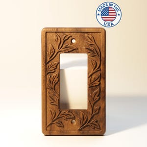 Peut inclure: Un cache-interrupteur en bois avec un motif floral sculpté. Le couvercle rectangulaire a une seule ouverture et est en bois brun foncé. Le motif présente des feuilles et des vignes sur les bords. Le texte "MADE IN THE USA" est dans un cercle en haut à droite.