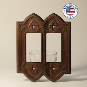 Peut inclure: Un cache-interrupteur en bois brun foncé avec un design en arc gothique. Le cache-interrupteur comporte deux ouvertures rectangulaires et est décoré de sculptures florales. Les mots "MADE IN THE USA" sont imprimés sur un autocollant circulaire.