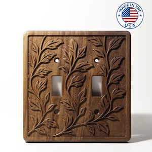 Peut inclure: Un cache-interrupteur double en bois avec un motif floral sculpté. Le couvercle est de couleur marron chaud et comporte deux ouvertures pour interrupteurs et deux trous de vis. Les mots "MADE IN THE USA" sont dans un cercle en haut à droite.
