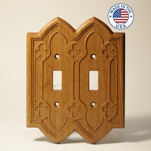 May include: Wooden double light switch cover with an ornate, gothic-style design. The cover is made of light brown wood and features two switch openings and decorative floral carvings. A "Made in the USA" sticker is in the upper right corner.