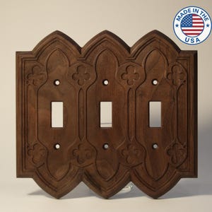 May include: A dark brown, wooden triple light switch cover with an ornate, gothic-style design. The cover features three switch openings and decorative floral accents. The words "MADE IN THE USA" are in a circle in the upper right corner.