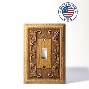 May include: A rectangular light switch cover plate featuring a wood grain finish and intricate carved leaf scrollwork patterns. The center includes a vertical toggle switch opening with two screw holes. A circular Made in the USA logo is visible.
