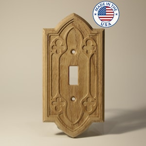 May include: A wooden light switch cover with an ornate, gothic-style design. The cover is made of light-colored wood and features carved floral and geometric patterns. The words "Made in the USA" are printed on a circular sticker in the upper right corner.
