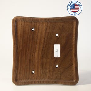 May include: A solid walnut wood light switch cover with a curved design. The cover has a single toggle switch opening and four screw holes. The words "Made in the USA" are in a circle in the upper right corner.