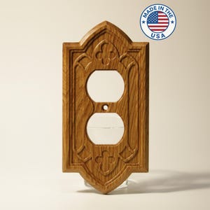 May include: A wooden electrical outlet cover with an ornate, carved design. The cover is made of light brown wood and features two circular openings for the outlets and a smaller opening for a screw. The design includes floral and cross-like elements. The words "MADE IN THE USA" are in a circle in the upper right corner.