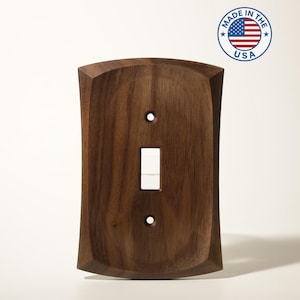 May include: A dark brown wooden light switch cover with a single switch opening and two screw holes. The cover has a curved top and bottom edge. A circular sticker with the text "MADE IN THE USA" and an American flag is in the upper right corner.