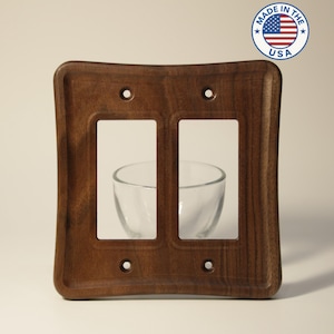 May include: A dark brown, wooden double light switch plate with a curved design. The plate has two rectangular openings and four screw holes. A "Made in the USA" sticker is in the upper right corner.