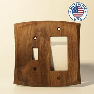 May include: A wooden light switch cover with a curved top and a rectangular opening for a toggle switch and a rocker switch. The cover is made of dark brown wood and has a natural wood grain pattern. The words "MADE IN THE USA" are in a circle with the American flag.