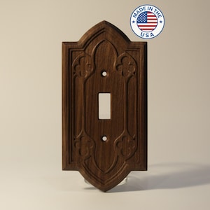 May include: A dark brown wooden light switch cover with an ornate, gothic-style design. The cover features a single switch opening and decorative floral carvings. A circular sticker with the American flag and the text "MADE IN THE USA" is in the upper right corner.