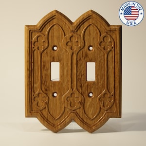 May include: A wooden double light switch cover with an ornate, carved design. The cover is made of light brown wood and features two switch openings and decorative floral-like patterns. The words "MADE IN THE USA" are printed on a circular sticker in the upper right corner.