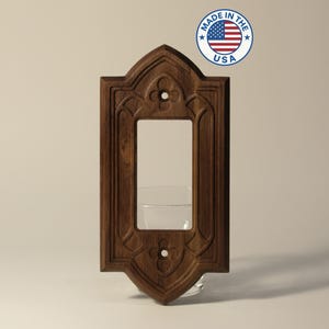 May include: A dark brown, wooden light switch cover with an ornate, gothic-style design. The cover has a rectangular opening for the switch and decorative carvings. The image includes a "Made in the USA" sticker.