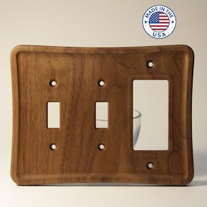 May include: A wooden light switch plate with a curved design. The plate has two toggle switch openings and one outlet opening. The plate is made of dark brown wood and has a smooth finish. The plate is marked "Made in the USA".