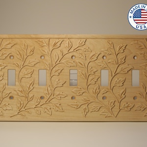 May include: A wooden light switch plate with a carved floral design. The plate has five switch openings and is made in the USA. The design features leaves and vines, creating a natural, decorative element.