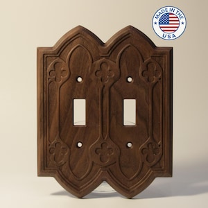 May include: A dark brown, wooden double light switch cover with an ornate, gothic-style design. The cover features two switch openings and decorative floral patterns. A circular sticker with the text "Made in the USA" is in the upper right corner.