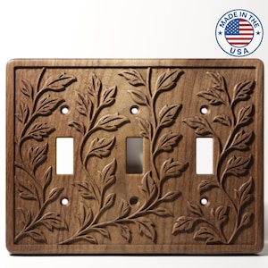 May include: A wooden light switch cover with a carved floral design. The cover has three switch openings and is made in the USA. The design features detailed leaves and vines, adding a natural aesthetic to any room.