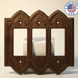 May include: A dark brown, wooden triple light switch cover with an ornate, gothic-style design. The cover features three rectangular openings and decorative floral accents. The product is marked "MADE IN THE USA".