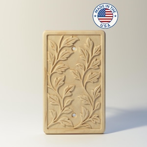 May include: A rectangular light-colored wooden light switch cover plate featuring a carved relief pattern of two intertwining leafy vine branches. The plate includes two circular screw holes and a circular Made in the USA emblem.