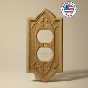 May include: A wooden light switch cover with a decorative carved design. The cover has two circular openings for outlets and is made in the USA. The design includes floral and geometric patterns.