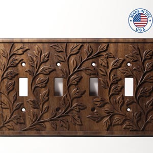 May include: A wooden light switch cover with a carved floral design. The cover has four screw holes and three switch openings. The design features leaves and vines. The words "MADE IN THE USA" are in a circle in the upper right corner.