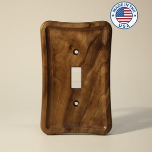May include: A wooden light switch cover with a natural wood grain finish. The cover has a rectangular shape with rounded edges and a recessed area around the switch opening. The switch is white. A "Made in the USA" sticker is in the upper right corner.