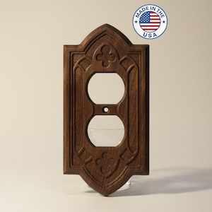 May include: A dark brown, wooden double outlet cover with an ornate, gothic-style design. The cover has two circular openings for the outlets and a central screw hole. The words "MADE IN THE USA" are printed on a circular sticker.