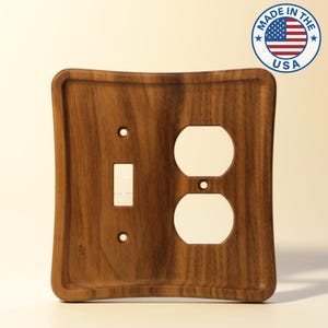 May include: A wooden light switch and outlet cover with a dark brown finish. The cover has a rectangular shape with curved edges and includes a switch opening, two outlet openings, and the text "MADE IN THE USA" in a circular emblem.