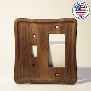 May include: A wooden light switch cover with a curved design. The cover has openings for a toggle switch and a rocker switch. The wood has a rich, dark brown color. A circular sticker with the text "MADE IN THE USA" is in the upper right corner.