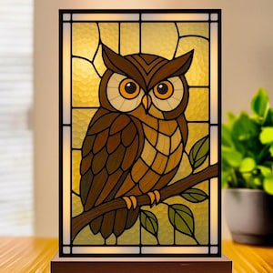 May include: A stained glass lamp featuring a brown owl perched on a branch. The owl has large orange eyes and is set against a yellow and gold background. The lamp is rectangular with a dark frame and a wooden base.